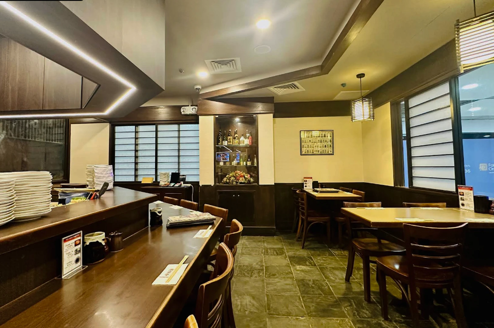 A wide shot of a traditional Japanese restaurant interior featuring dark wood accents and warm yellow lighting. On the left, a dark wooden counter with several stacks of white plates faces an open kitchen area. To the right, several small wooden dining tables with matching chairs are arranged on a dark, slate-tiled floor. A recessed shelf in the center wall displays various sake bottles, and a framed piece of art hangs on the back wall near a window with traditional horizontal slats.