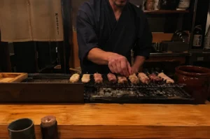 A medium shot captured from a low angle shows a chef in a dark blue, traditional Japanese garment meticulously arranging a row of chicken skewers on a rectangular charcoal grill. The chef, seen from the chest down, focuses on the variety of yakitori—ranging from pale, round pieces to darker, more textured cuts—which are neatly aligned over glowing embers and white ash. The setting is a dimly lit, rustic izakaya with warm wooden countertops in the foreground and shelves holding pottery and glass bottles in the background. A brown ceramic jar and a small container of toothpicks sit on the polished wood, adding to the intimate, authentic atmosphere of the kitchen.