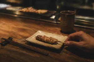 A minimalist dining moment at a traditional Japanese yakitori bar. In the center, a single skewer of perfectly charred, golden-brown grilled chicken (yakitori) sits on an artisanal, cream-colored ceramic plate with organic edges. A hand enters from the right, fingers gently grasping the wooden end of the skewer, suggesting a personal and candid interaction with the food. To the left, dark wooden chopsticks rest on a matching black rest, and in the background, a rustic, textured ceramic tea cup stands beside a glass partition that reveals the soft, blurred glow of more skewers grilling over a charcoal fire. The scene is illuminated by warm, low-key lighting that emphasizes the rich textures of the weathered wooden countertop and the glistening juices of the meat, evoking a quiet, sophisticated, and authentic culinary atmosphere.