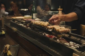 A yakitori chef skillfully tending to rows of chicken skewers over a traditional narrow charcoal grill in a dimly lit, upscale izakaya. The chef’s hand is in motion, turning a skewer amidst a light plume of smoke rising from the glowing embers of white Binchotan charcoal nestled in the grill’s trough. In the blurred background, several patrons watch the process from across a dark wooden counter, while a green bottle of sake sits nearby. In the bottom-left foreground, a finished skewer accompanied by a lemon wedge rests on a slate-colored ceramic plate, emphasizing the contrast between the raw heat of the grill and the refined presentation of the meal.
