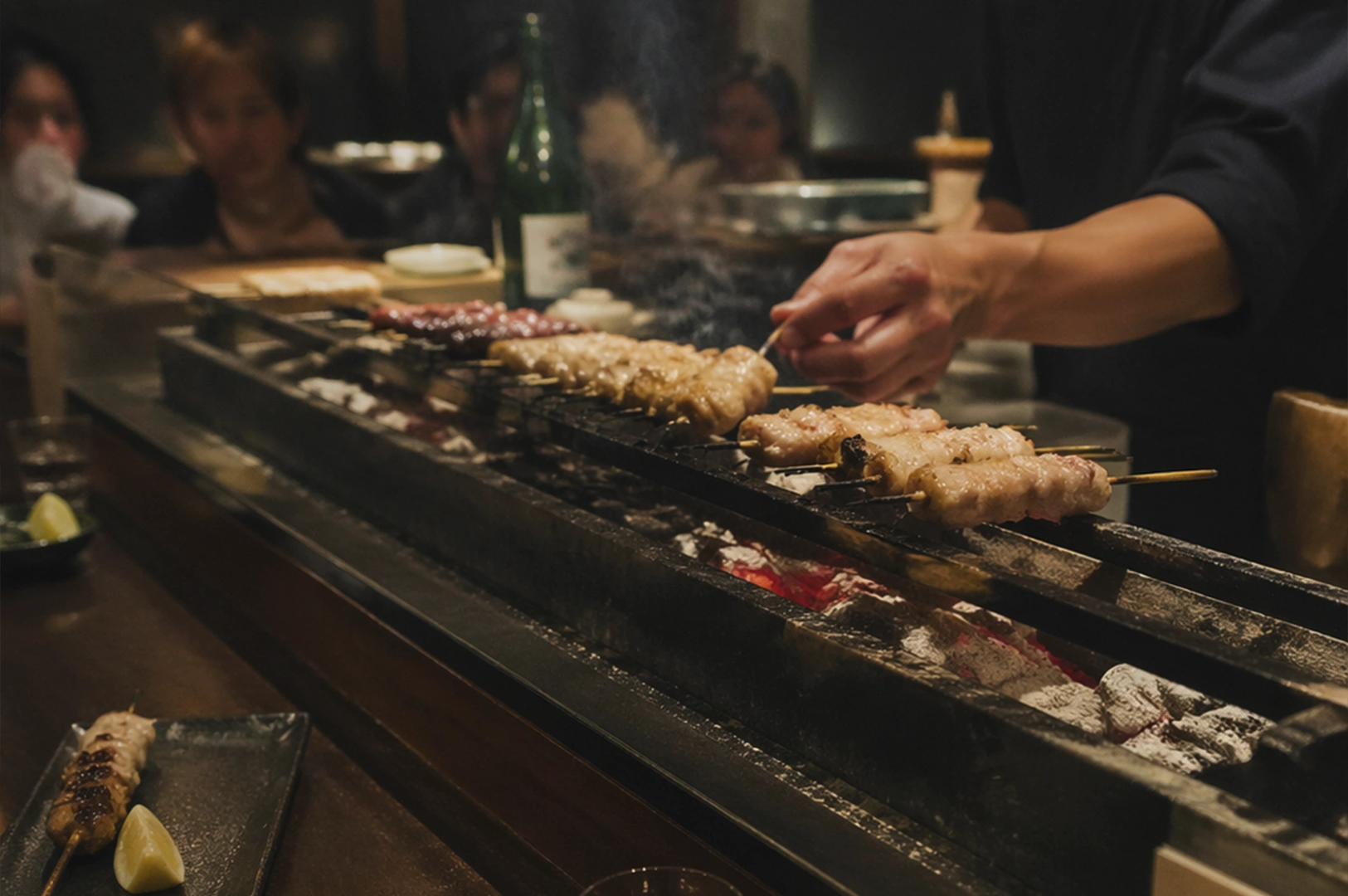 A yakitori chef skillfully tending to rows of chicken skewers over a traditional narrow charcoal grill in a dimly lit, upscale izakaya. The chef’s hand is in motion, turning a skewer amidst a light plume of smoke rising from the glowing embers of white Binchotan charcoal nestled in the grill’s trough. In the blurred background, several patrons watch the process from across a dark wooden counter, while a green bottle of sake sits nearby. In the bottom-left foreground, a finished skewer accompanied by a lemon wedge rests on a slate-colored ceramic plate, emphasizing the contrast between the raw heat of the grill and the refined presentation of the meal.