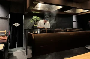 A Japanese yakitori chef in a white uniform meticulously grilling skewers over a charcoal fire in a modern, upscale restaurant setting with a bonsai tree in the background.