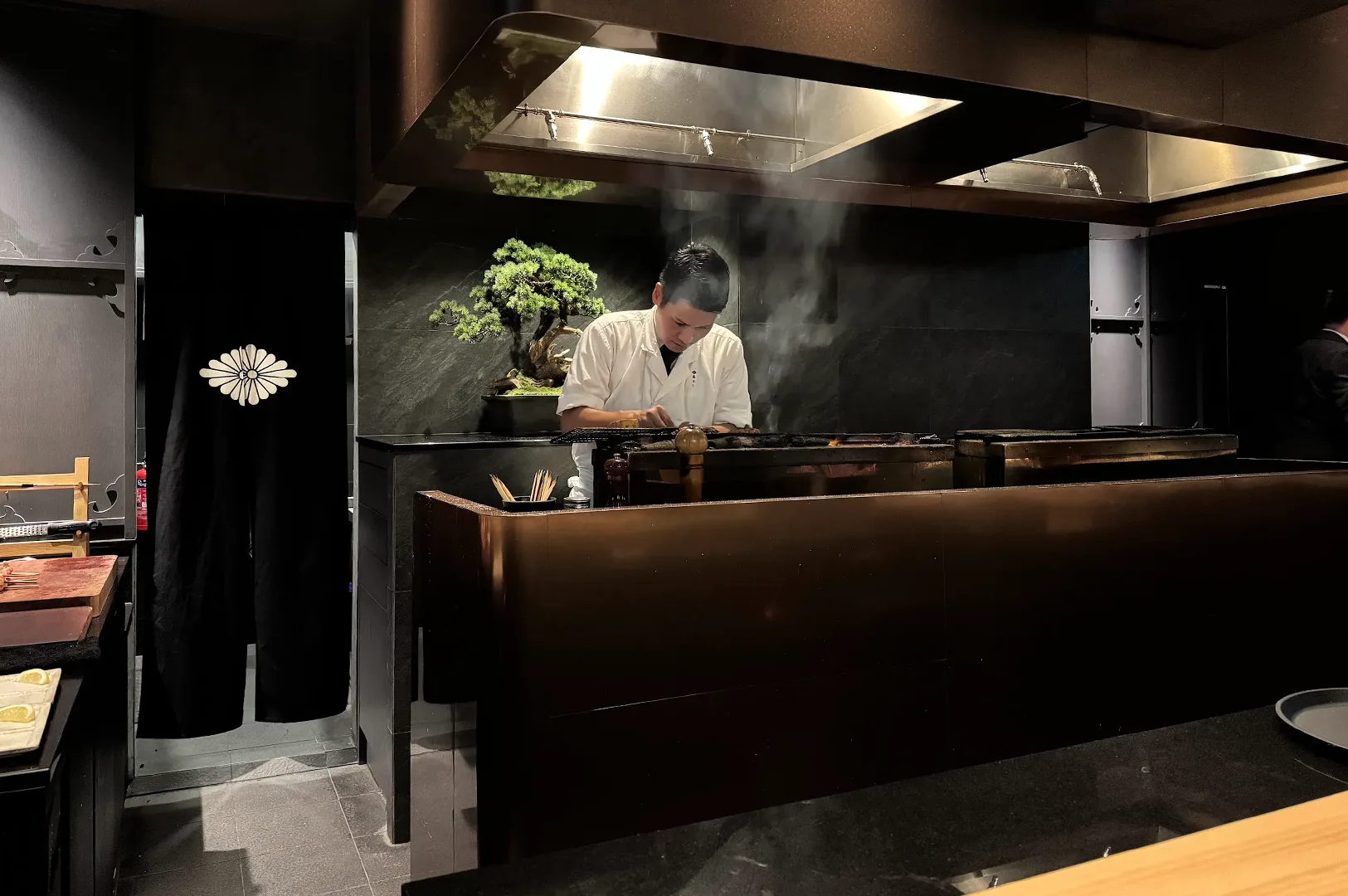 A Japanese yakitori chef in a white uniform meticulously grilling skewers over a charcoal fire in a modern, upscale restaurant setting with a bonsai tree in the background.