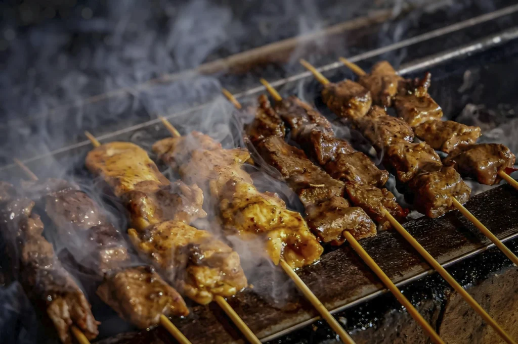 A close-up, high-angle shot of several meat skewers (yakitori) cooking over a charcoal grill. Wisps of white smoke rise between the rows of skewers. The meat on the left appears to be chicken with a light glaze, while the skewers on the right are darker, more charred, and glistening with sauce.