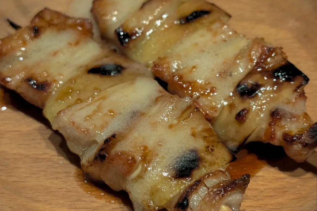 An extreme close-up of two Japanese yakitori skewers resting on a light-colored wooden plate. The skewers feature thick, succulent pieces of grilled meat—likely pork belly or chicken—with a glistening, caramelized glaze. The meat shows distinct charred grill marks and rendered fat, indicating it was cooked over an open flame. The lighting is warm, highlighting the texture and moisture of the food.