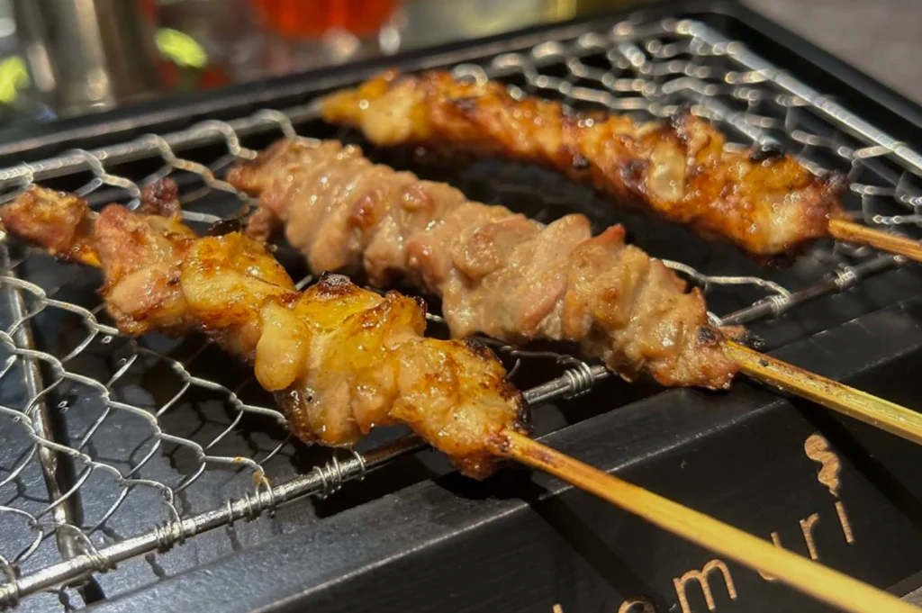 A high-angle, close-up shot of three yakitori skewers resting on a black tabletop grill with a wire mesh top. The chicken pieces are golden-brown and glistening with glaze, showing slight char marks from the charcoal. The grill is embossed with the "kemuri" logo in gold lettering. The background is softly blurred, showing hints of glassware and warm ambient lighting