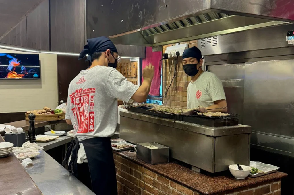 Two male chefs wearing black face masks, black headwraps, and light grey t-shirts work behind a long yakitori grill. The chef in the foreground has his back to the camera, revealing a red "40 Years Anniversary" graphic on his shirt. The chef in the background is carefully attending to numerous skewers of meat and seafood cooking over an open flame. The grill area is built into a rustic brick base under a large stainless steel industrial exhaust hood.