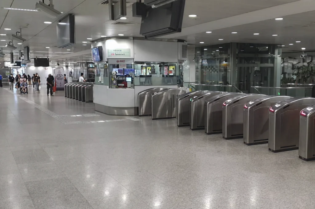 A wide-angle, eye-level shot of a modern, brightly lit train station concourse, Somerset MRT in Singapore. Two long rows of sleek, stainless steel ticket gantries with glowing red indicators lead toward a white passenger service office in the center. The floors are polished grey granite, reflecting the overhead fluorescent lights. In the background, several commuters wearing face masks walk through the spacious, clean terminal, which features glass-walled elevators and digital display screens hanging from the ceiling.
