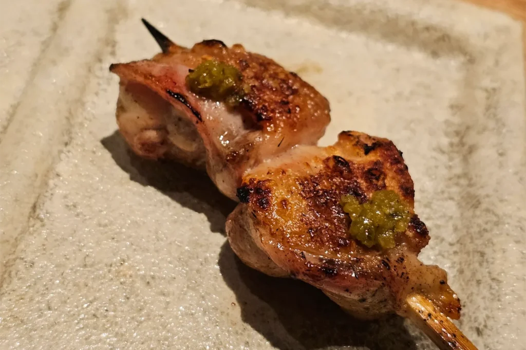 Close-up of a perfectly grilled chicken yakitori skewer topped with green yuzu kosho paste, served on a textured ceramic plate.