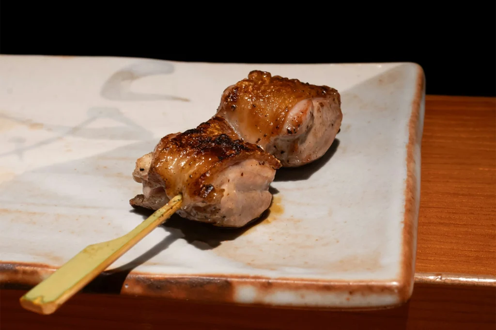 A single yakitori skewer rests on a rustic, rectangular ceramic plate with a speckled white and brown glaze. The skewer features two thick pieces of grilled chicken with perfectly browned, crispy skin. The background is a minimalist dark void, highlighting the texture of the meat and the wooden table surface below.