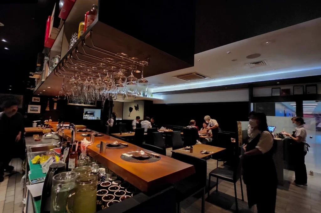 A wide-angle interior view of Kazu Sumiyaki Restaurant showing a long, polished wooden bar counter in the foreground. Upside-down wine glasses hang from a rack above the bar, and several place settings with dark ceramic bowls are ready for guests. In the background, the dimly lit dining room features several patrons seated at dark tables. Staff members wearing black aprons and face masks are seen attending to customers and standing near a bright glass entrance on the right.