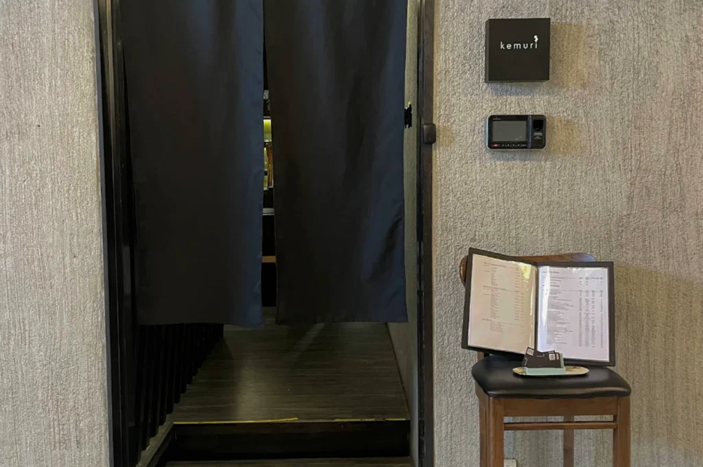 The minimalist entrance of a Japanese restaurant. Dark navy blue noren curtains hang over a doorway, partially obscuring a glimpse of the warm interior and wooden flooring. To the right, a textured grey concrete wall holds a small black square sign with the word "kemuri" in white script. Below the sign is a digital keypad and fingerprint scanner. A wooden chair sits in the foreground, holding an open menu and a small tray with business cards.