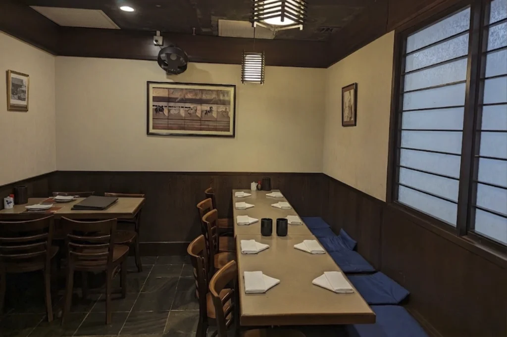 A cozy, dimly lit corner of the restaurant featuring a long wooden table set for six people. Each place setting includes a white napkin folded into a triangle. Along the wall, a long blue cushioned bench serves as seating, while three wooden chairs are tucked into the opposite side. The walls are cream-colored with dark wood wainscoting, decorated with two framed pieces of Japanese-style artwork and a traditional hanging lantern.