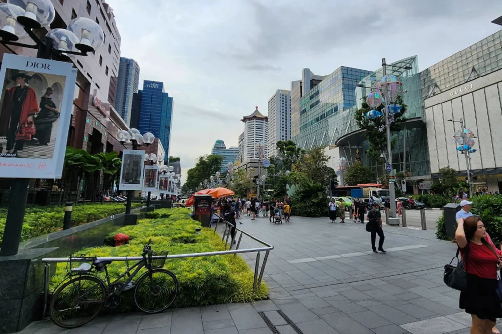 A bustling outdoor street scene on a wide, grey-tiled pedestrian walkway in a high-end shopping district. On the right, the glass facade of an ION Orchard mall is visible, featuring a large "GUCCI" storefront. To the left, large Dior fashion advertisements are mounted on lampposts beside lush green landscaping. A bicycle is parked in the foreground against a metal railing. In the distance, the iconic Pagoda-style roof of the Tang Plaza stands out against a cloudy sky, while various shoppers and pedestrians navigate the urban space.