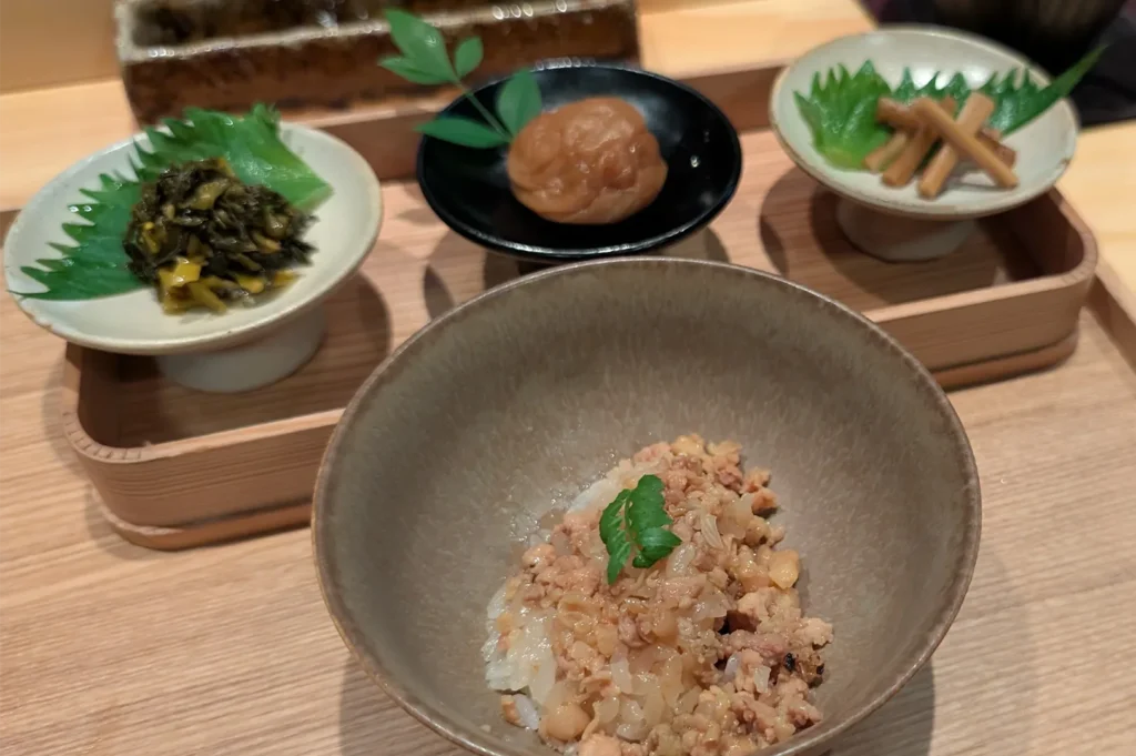 A traditional Japanese meal set featuring a bowl of minced chicken over rice (donabe rice) accompanied by small side dishes of pickles, umeboshi, and burdock root.