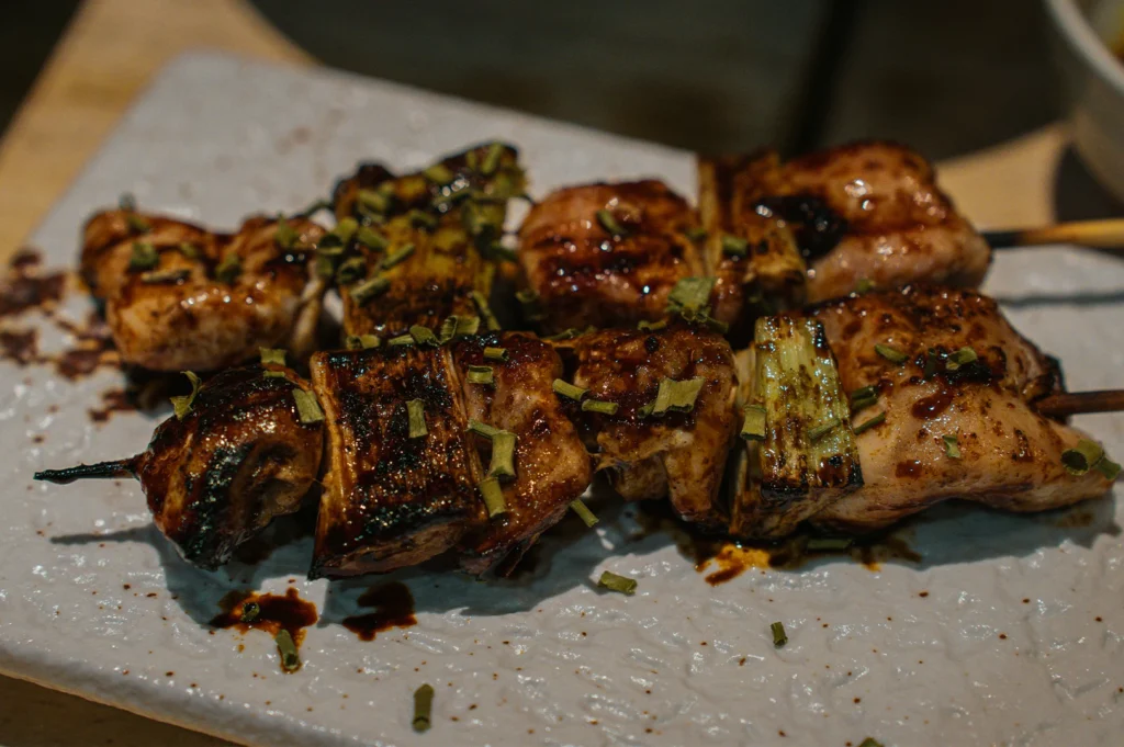 Two yakitori skewers are presented on a textured white ceramic plate. The skewers consist of alternating pieces of grilled chicken and thick scallion rounds, all heavily coated in a dark, glossy savory sauce and garnished with chopped green herbs. Stray drops of sauce and herbs are scattered across the plate.
