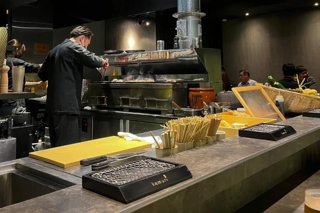 Dining counter looking into the active kitchen. A chef in a black uniform is seen from the back, using tongs to turn skewers over a long, glowing charcoal hearth. The counter in the foreground features bundles of bamboo skewers, a yellow prep board, and a small personal grill. In the background, patrons are seated at the far end of the counter, contributing to the busy, atmospheric dining environment.