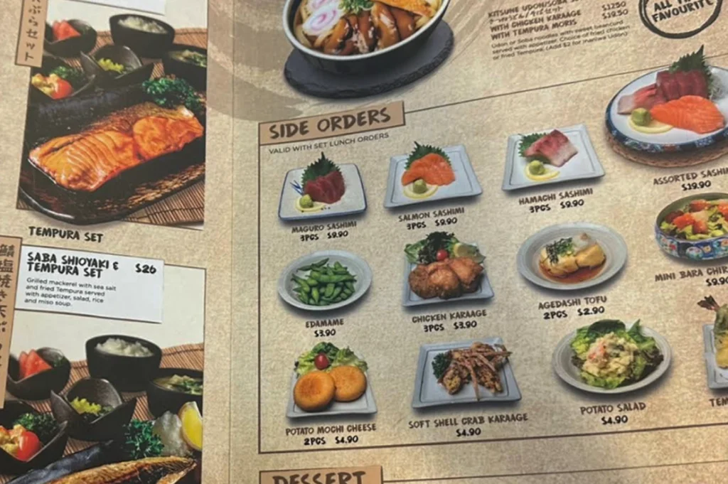 A high-angle shot of a printed physical menu highlighting "Side Orders" and "Set Lunch" options. The side orders include Maguro Sashimi, Salmon Sashimi, and Hamachi Sashimi for $9.90 each, alongside Edamame, Chicken Karaage, Agedashi Tofu, and Potato Mochi Cheese. To the left, a large photo of a "Saba Shioyaki & Tempura Set" is priced at $26. The layout is clean with clear photos of each dish on a textured, light-brown background.