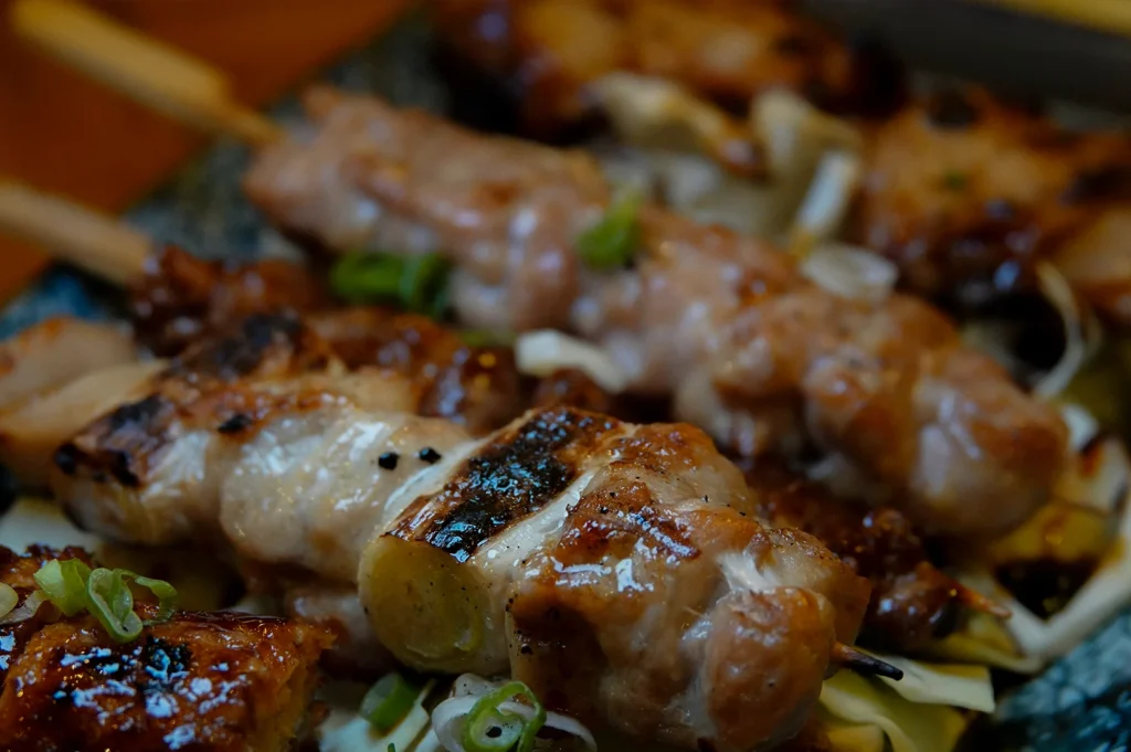 An extreme close-up, shallow-depth-of-field shot of several grilled chicken skewers. The focus is sharp on a piece of charred scallion and juicy chicken in the foreground. The meat is glistening with oil and juices, garnished with thinly sliced green onions. Other skewers are visible but softly blurred in the background.