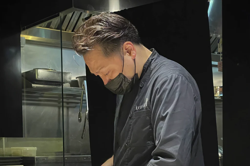 Profile of a chef in a dark kitchen. The chef wears a black face mask and a black uniform with "kemuri" embroidered on the chest pocket. He is looking down with deep concentration, his hair styled with a slight wet-look sheen. Harsh, dramatic overhead lighting catches the top of his head and shoulders, contrasting sharply with the pitch-black background and the stainless steel kitchen equipment visible in the soft-focus behind him.