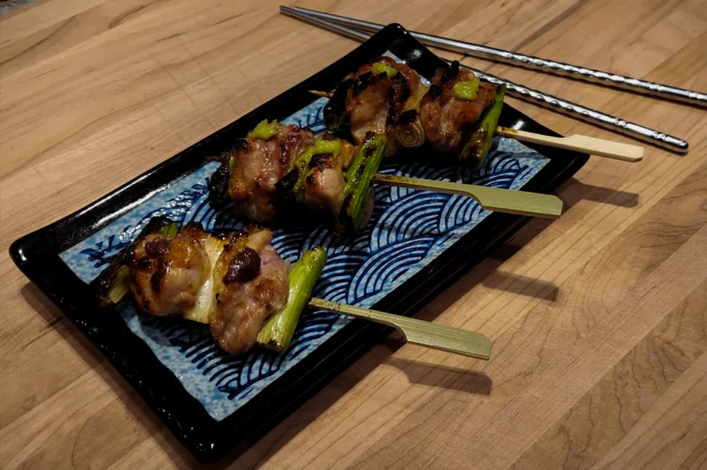 Three yakitori skewers are arranged diagonally on a black rectangular plate with a blue and white "Seigaiha" (wave) pattern. Each skewer has grilled chicken and green onions, topped with a small dollop of bright green wasabi. A pair of textured metal chopsticks rests on the light wood grain table behind the plate.