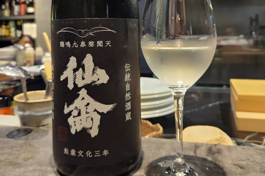 A bottle of Japanese sake next to a filled glass. The bottle has a matte black label featuring bold, silver-white kanji characters and smaller traditional text. Next to it, a delicate, long-stemmed wine glass contains a slightly cloudy, pale white sake. The setting is a textured grey dining counter, with blurred stacks of white ceramic plates and kitchen items in the background under warm, spot-lit ambiance.