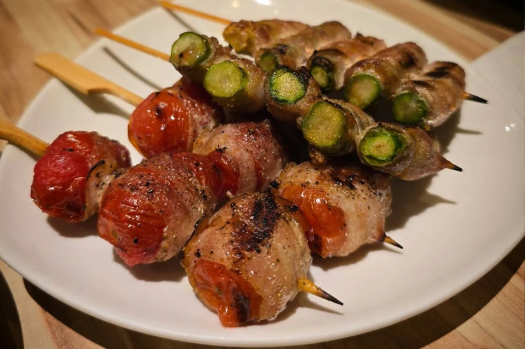 A close-up, top-down shot of a white plate holding several skewers of grilled yakitori. In the foreground are four skewers of bright red cherry tomatoes wrapped in crispy, seasoned bacon. Tucked behind them are several skewers of green asparagus, also wrapped in thin slices of grilled pork or bacon. The food is glistening, showing char marks from the grill and a light dusting of black pepper.