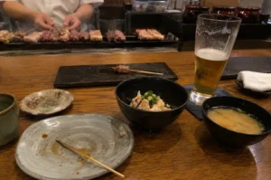 A high-angle, eye-level shot captures a person's perspective at a wooden yakitori counter, signifying the end of a meal. In the foreground, a large, empty gray ceramic plate sits with a single used wooden skewer and sauce residue, flanked by a small bowl of miso soup and a bowl of rice topped with minced meat and scallions. A tall, half-empty glass of light amber beer sits on a blue coaster to the right. In the mid-ground, a black rectangular serving slate holds one final lonely skewer. The background, slightly out of focus, shows a chef’s hands expertly tending to several rows of chicken skewers grilling over a smoky charcoal hearth, while dark ceramic jars and kitchen containers line the back shelf. The warm, ambient lighting and the scattered remains of the meal create an intimate, satisfied atmosphere of a traditional Japanese dining experience.