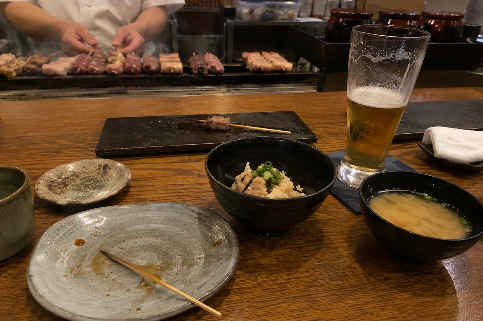 A high-angle, eye-level shot captures a person's perspective at a wooden yakitori counter, signifying the end of a meal. In the foreground, a large, empty gray ceramic plate sits with a single used wooden skewer and sauce residue, flanked by a small bowl of miso soup and a bowl of rice topped with minced meat and scallions. A tall, half-empty glass of light amber beer sits on a blue coaster to the right. In the mid-ground, a black rectangular serving slate holds one final lonely skewer. The background, slightly out of focus, shows a chef’s hands expertly tending to several rows of chicken skewers grilling over a smoky charcoal hearth, while dark ceramic jars and kitchen containers line the back shelf. The warm, ambient lighting and the scattered remains of the meal create an intimate, satisfied atmosphere of a traditional Japanese dining experience.