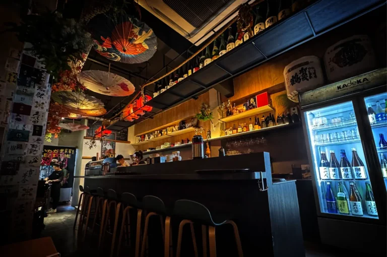 A wide, low-angle shot of a moody Japanese izakaya bar with warm, ambient lighting. A long, dark counter lined with black barstools runs through the center. Behind it, shelves are stocked with liquor and sake bottles, flanked by a glowing glass-door beverage cooler on the right displaying chilled sake. The ceiling is decorated with traditional elements, including colorful hanging paper umbrellas, glowing red lanterns, and large sake barrels resting on an upper ledge.