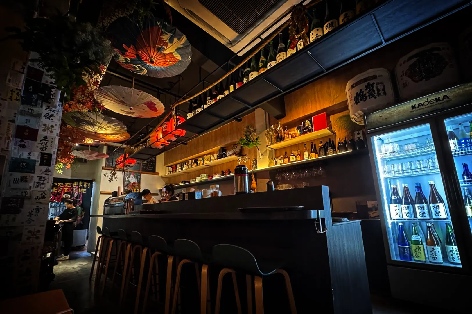 A wide, low-angle shot of a moody Japanese izakaya bar with warm, ambient lighting. A long, dark counter lined with black barstools runs through the center. Behind it, shelves are stocked with liquor and sake bottles, flanked by a glowing glass-door beverage cooler on the right displaying chilled sake. The ceiling is decorated with traditional elements, including colorful hanging paper umbrellas, glowing red lanterns, and large sake barrels resting on an upper ledge.