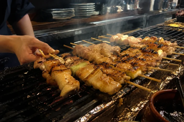 A close-up, eye-level shot captures a chef’s left hand carefully turning wooden skewers of chicken yakitori on a traditional, long, narrow Japanese charcoal grill. Rows of various chicken skewers, including negima (chicken thighs alternated with thick pieces of Japanese leek), are sizzling over the hot grates, showing prominent grill marks, charred edges, and a glistening glaze. In the background, wisps of white smoke rise from the grill toward a dimly lit, professional izakaya kitchen setting featuring stacked ceramic plates on a counter, while a small brown ceramic pot containing dark tare sauce sits in the bottom right corner.