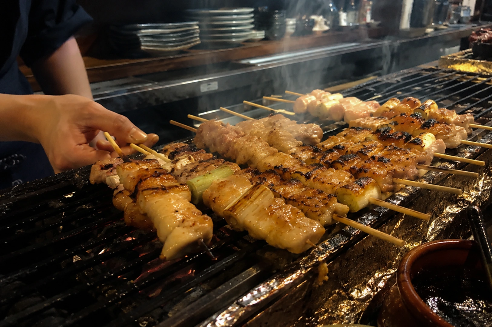 A close-up, eye-level shot captures a chef’s left hand carefully turning wooden skewers of chicken yakitori on a traditional, long, narrow Japanese charcoal grill. Rows of various chicken skewers, including negima (chicken thighs alternated with thick pieces of Japanese leek), are sizzling over the hot grates, showing prominent grill marks, charred edges, and a glistening glaze. In the background, wisps of white smoke rise from the grill toward a dimly lit, professional izakaya kitchen setting featuring stacked ceramic plates on a counter, while a small brown ceramic pot containing dark tare sauce sits in the bottom right corner.