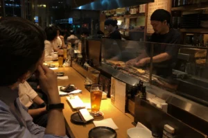 A wide-angle, eye-level shot from a customer's perspective at a long, light-wood yakitori counter. In the foreground, the back of a man’s head and shoulder are visible as he rests his chin on his hand, looking toward the grill. Two chefs in black uniforms and head wraps are working behind a glass partition, tending to dozens of chicken skewers over a long charcoal grill. A tall glass of amber-colored beer with condensation sits prominently on the counter, surrounded by small plates, chopsticks, and white napkins. The background shows other patrons seated along the counter and the warm, bustling interior of the restaurant at night.