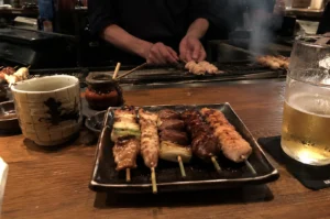 A close-up, eye-level shot of a rectangular ceramic plate holding five diverse yakitori skewers on a wooden bar counter. The skewers include glazed chicken, scallions, and minced meat. In the background, a chef’s hands are visible over a steaming charcoal grill, while a frosted glass of beer and a traditional ceramic tea cup flank the plate.
