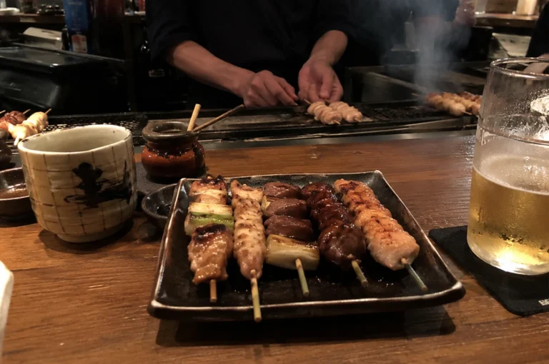 A close-up, eye-level shot of a rectangular ceramic plate holding five diverse yakitori skewers on a wooden bar counter. The skewers include glazed chicken, scallions, and minced meat. In the background, a chef’s hands are visible over a steaming charcoal grill, while a frosted glass of beer and a traditional ceramic tea cup flank the plate.