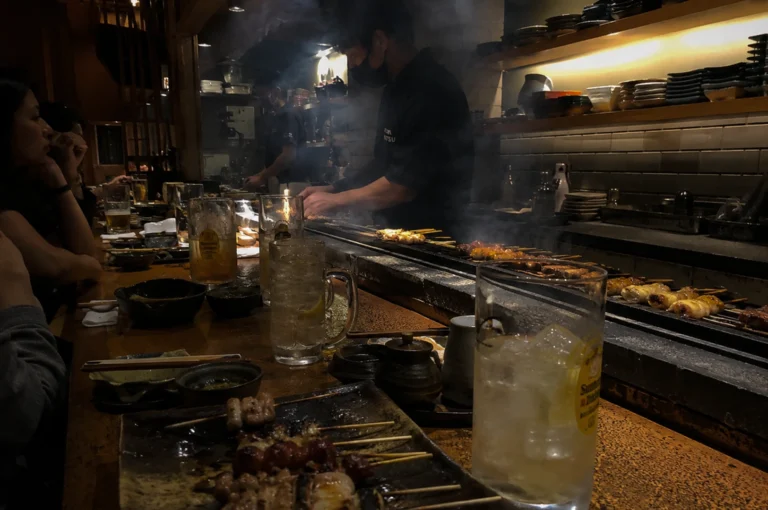 An intimate, dimly lit yakitori restaurant features a long wooden counter where customers sit watching a chef grill numerous skewers of meat over a smoky, built-in counter-top grill. In the foreground, a close-up shows a tall glass of an iced beverage, a plate holding freshly grilled skewers, and small condiment dishes on the dark wood surface. Behind the counter, the chef in a black uniform and face mask focuses on flipping skewers on the long, narrow grill, which is illuminated by a small flame and filled with rising white smoke. Shelves stacked with ceramic plates and kitchenware line the tiled wall in the warm, atmospheric background, while patrons converse softly on the left side of the frame.