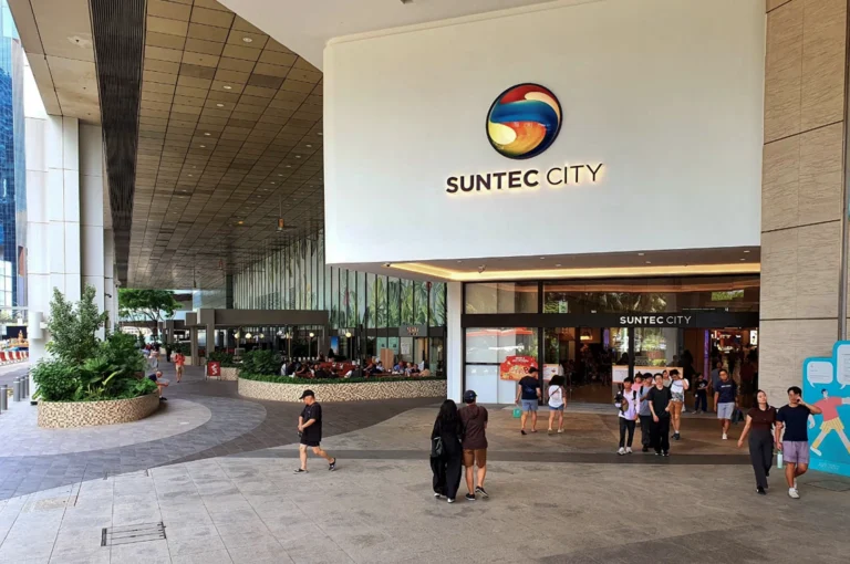 An outdoor wide shot of the Suntec City mall entrance under a large white overhang. The "SUNTEC CITY" logo is prominently displayed in gold lettering beneath a circular multi-colored emblem. People in casual summer attire walk across the light grey tiled plaza. To the left, there is a recessed dining area with greenery in stone planters, and the architectural lines of the building's pillars and ceiling create a strong sense of depth leading toward the street.