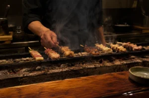 A chef in a dark uniform meticulously tends to a row of yakitori skewers grilling over a traditional narrow charcoal bed, known as a konro. Thick plumes of white smoke rise from the sizzling meat, partially obscuring the chef’s torso and adding a sense of movement to the dimly lit, atmospheric kitchen. The skewers feature various cuts of chicken and green scallions, some showing a deep, caramelized char while others are still pale and cooking. In the foreground, the edge of a polished wooden counter and a small ceramic dish are visible, while the background reveals the organized clutter of a professional Japanese kitchen with stacked bowls and stainless steel surfaces.