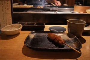 A single skewer of glistening, glazed yakitori sits centered on a dark, textured ceramic plate atop a light-colored wooden counter in a dimly lit, intimate Japanese restaurant. The skewer, likely chicken or meatballs, has a rich amber sheen and rests on a plate with traditional ribbed patterns. Surrounding the plate are various dining essentials, including a small stoneware bowl, a three-compartment condiment dish, and a ceramic cup filled with tea. In the soft-focus background, the hands of a chef are visible behind a raised counter, working over a smoky grill where more skewers are being prepared, while a handwritten Japanese menu stands partially visible on the left, adding to the authentic atmosphere.