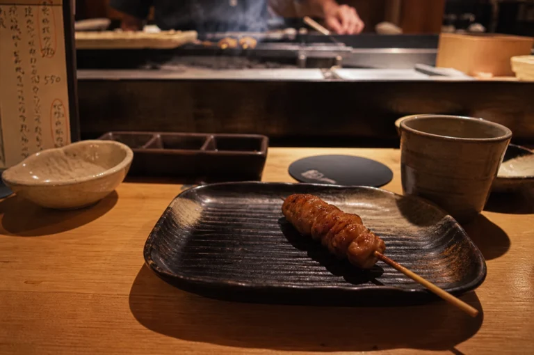 A single skewer of glistening, glazed yakitori sits centered on a dark, textured ceramic plate atop a light-colored wooden counter in a dimly lit, intimate Japanese restaurant. The skewer, likely chicken or meatballs, has a rich amber sheen and rests on a plate with traditional ribbed patterns. Surrounding the plate are various dining essentials, including a small stoneware bowl, a three-compartment condiment dish, and a ceramic cup filled with tea. In the soft-focus background, the hands of a chef are visible behind a raised counter, working over a smoky grill where more skewers are being prepared, while a handwritten Japanese menu stands partially visible on the left, adding to the authentic atmosphere.
