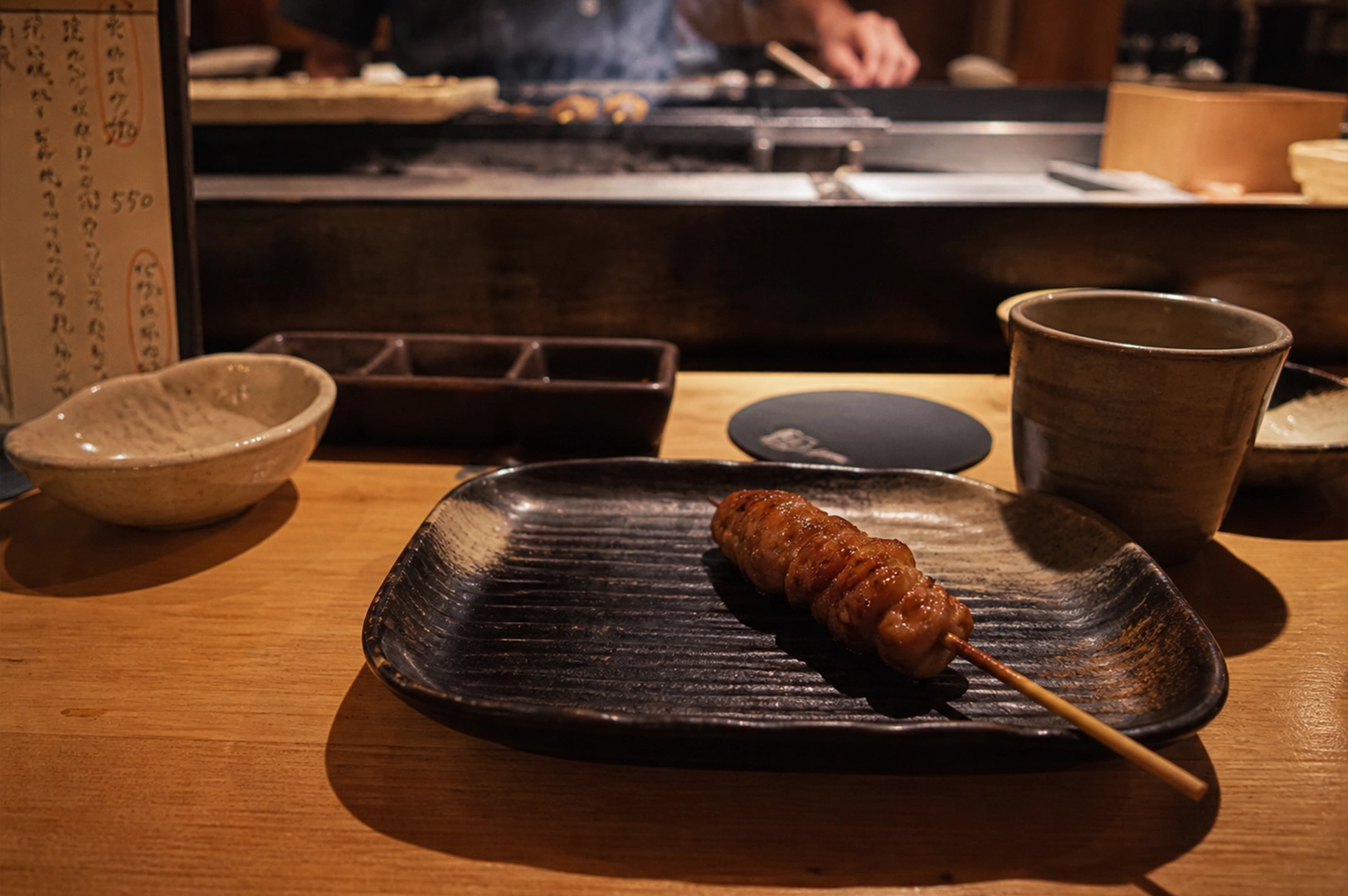 A single skewer of glistening, glazed yakitori sits centered on a dark, textured ceramic plate atop a light-colored wooden counter in a dimly lit, intimate Japanese restaurant. The skewer, likely chicken or meatballs, has a rich amber sheen and rests on a plate with traditional ribbed patterns. Surrounding the plate are various dining essentials, including a small stoneware bowl, a three-compartment condiment dish, and a ceramic cup filled with tea. In the soft-focus background, the hands of a chef are visible behind a raised counter, working over a smoky grill where more skewers are being prepared, while a handwritten Japanese menu stands partially visible on the left, adding to the authentic atmosphere.