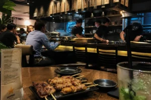A wide-angle view from a diner's perspective inside a dimly lit, atmospheric Japanese restaurant. In the foreground, a dark plate of glistening grilled skewers sits on a wooden table next to a tall glass of a green cocktail with mint leaves. In the background, three chefs are visible behind a long wooden counter, working in an open kitchen where steam rises from the cooking area. Customers sit at the counter and at nearby tables, creating a warm, bustling social environment.