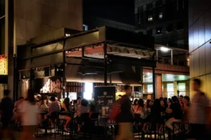 Outdoor dining area at a modern restaurant at night, with groups of people seated at tables under warm lighting. The black, open-air structure is surrounded by tall city buildings, while blurred pedestrians pass by in the foreground, creating a lively urban atmosphere.