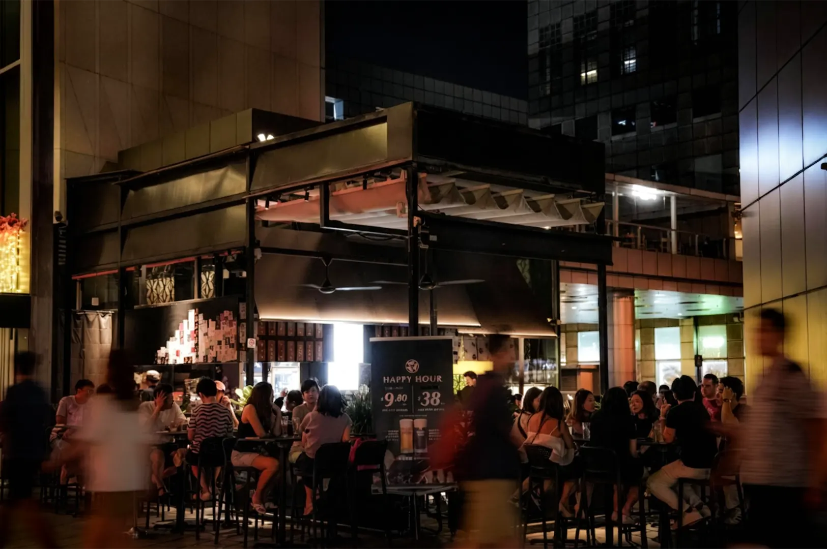 Outdoor dining area at a modern restaurant at night, with groups of people seated at tables under warm lighting. The black, open-air structure is surrounded by tall city buildings, while blurred pedestrians pass by in the foreground, creating a lively urban atmosphere.