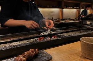 An angled, eye-level shot from the perspective of a diner seated at a light wood counter. In the foreground, a finished skewer sits on a dark ceramic plate next to a rustic tea cup. In the mid-ground, the chef's hands are visible through a thin veil of smoke, tending to skewers over a bed of glowing red coals. The background shows a secondary chef working in a deep, narrow kitchen lined with minimalist wooden cabinetry.