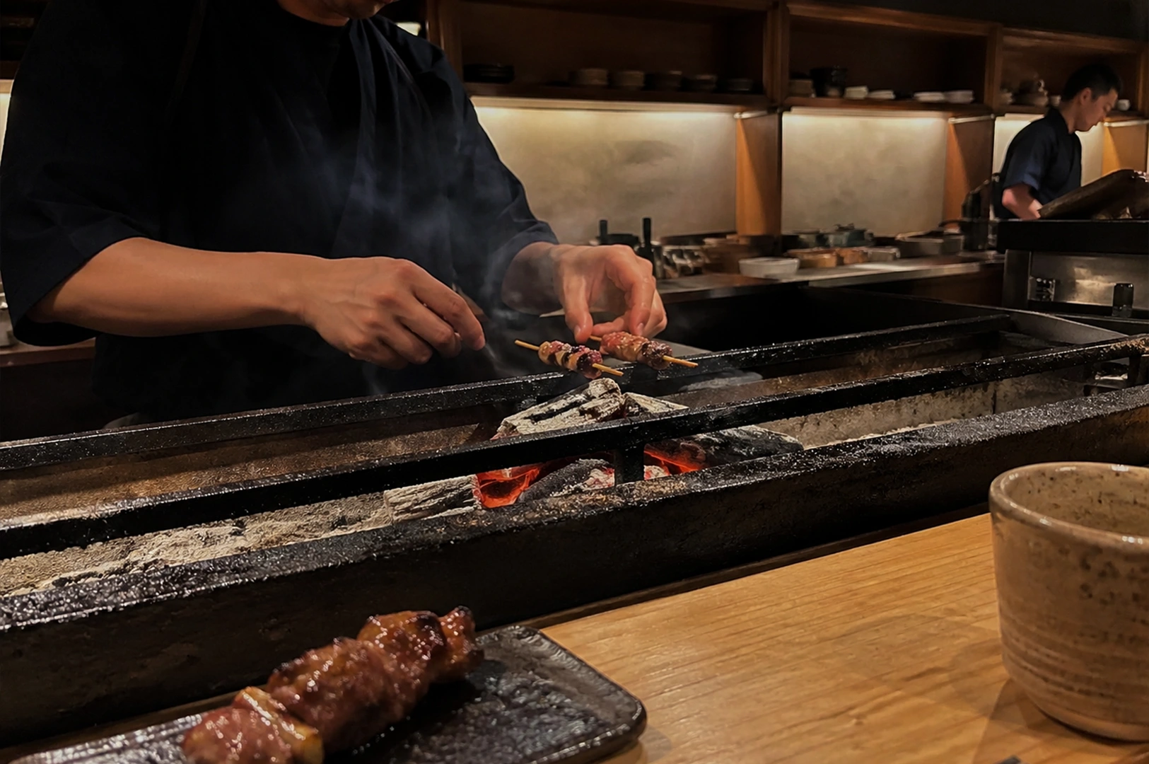 An angled, eye-level shot from the perspective of a diner seated at a light wood counter. In the foreground, a finished skewer sits on a dark ceramic plate next to a rustic tea cup. In the mid-ground, the chef's hands are visible through a thin veil of smoke, tending to skewers over a bed of glowing red coals. The background shows a secondary chef working in a deep, narrow kitchen lined with minimalist wooden cabinetry.