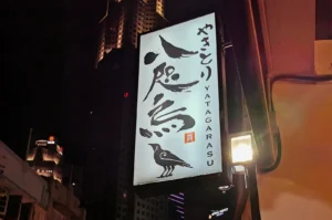 A lit-up outdoor signboard for Yakitori Yatagarasu restaurant at night featuring Japanese calligraphy and a three-legged crow logo with city buildings in the background.