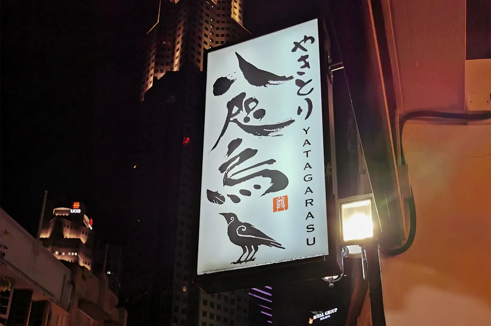 A lit-up outdoor signboard for Yakitori Yatagarasu restaurant at night featuring Japanese calligraphy and a three-legged crow logo with city buildings in the background.