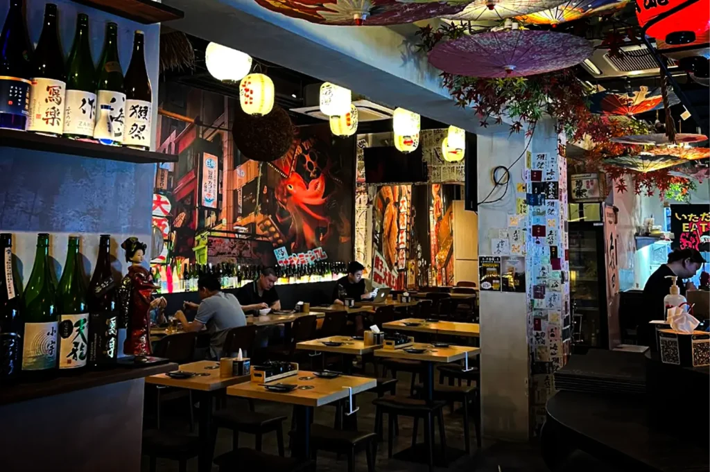 An eye-level shot of a vibrant, bustling Japanese izakaya dining room filled with small wooden tables and black chairs. Patrons are seated along the back wall, which is adorned with a colorful, lit-up mural depicting a Tokyo street scene at night. The restaurant is richly decorated with traditional elements, including rows of sake bottles on shelves in the foreground, hanging white and yellow paper lanterns, and decorative paper umbrellas suspended from the ceiling. A central support pillar is heavily covered in small, white paper slips.